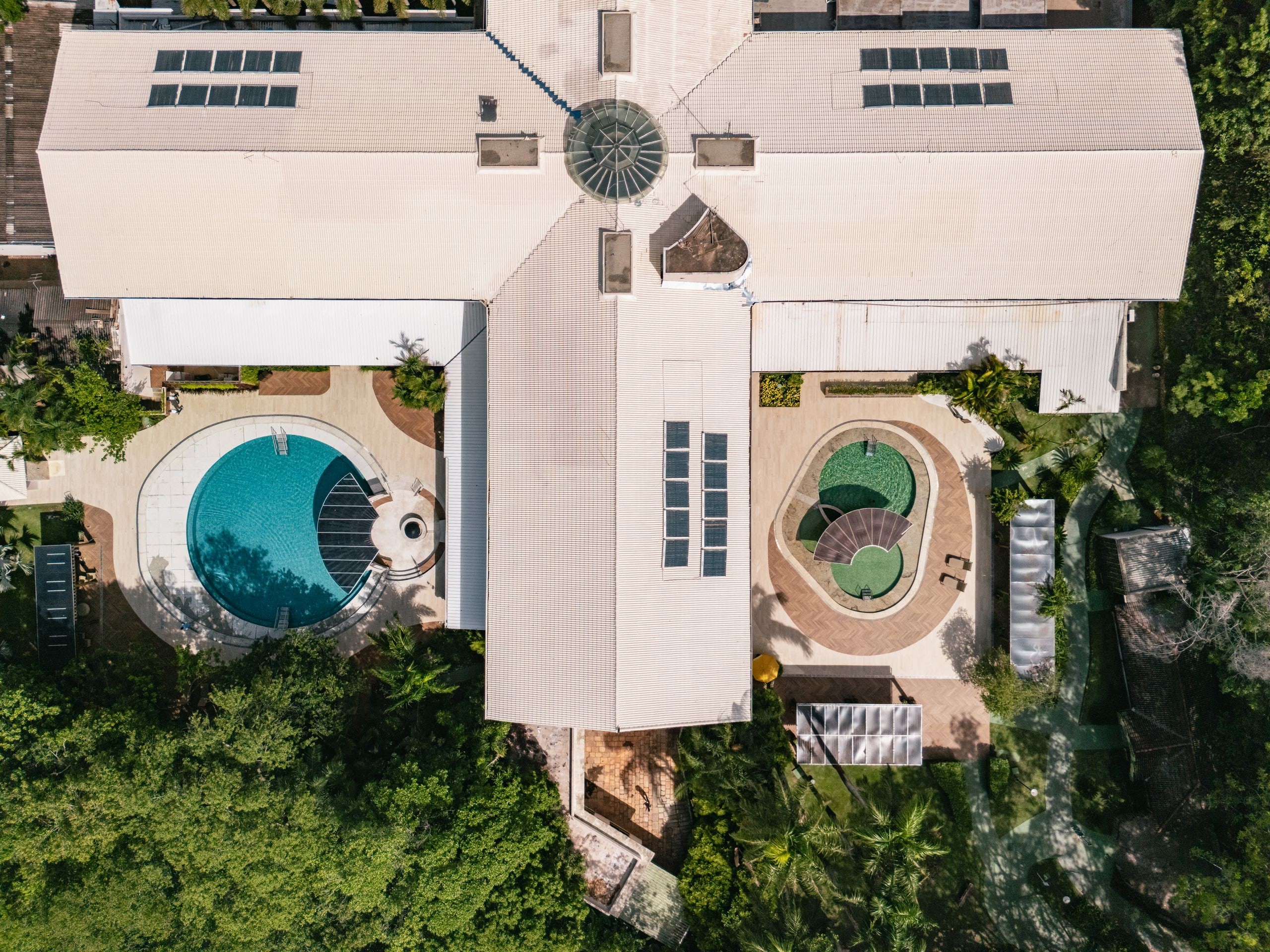 Vista a&eacute;rea de um complexo de lazer com telhados claros, duas piscinas e muita vegeta&ccedil;&atilde;o. &Agrave; esquerda, uma piscina azul em formato de C com deck de madeira. &Agrave; direita, duas piscinas menores de formato oval com &aacute;gua esverdeada. Pain&eacute;is solares e claraboias no telhado complementam a estrutura.