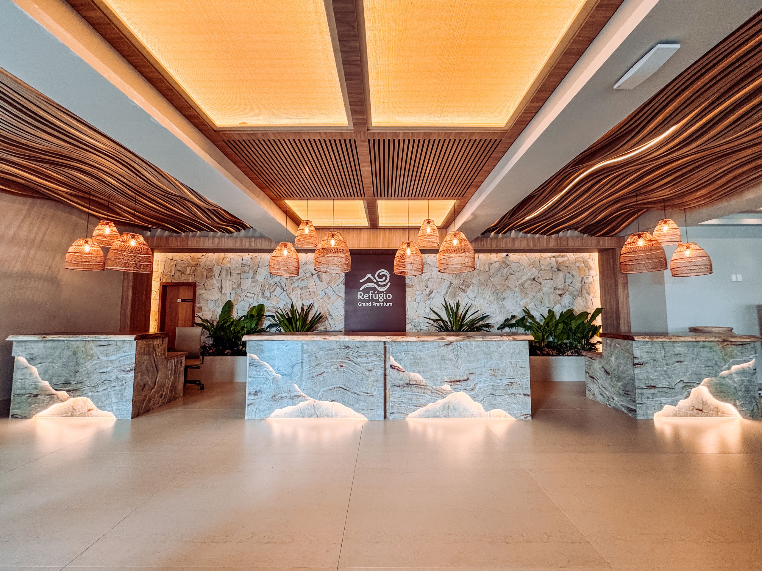 Recep&ccedil;&atilde;o de hotel com balc&otilde;es de m&aacute;rmore iluminados, teto de madeira ondulada e lumin&aacute;rias de palha penduradas. Ao fundo, uma parede de pedra com plantas verdes e o logo Ref&uacute;gio Grand Premium
