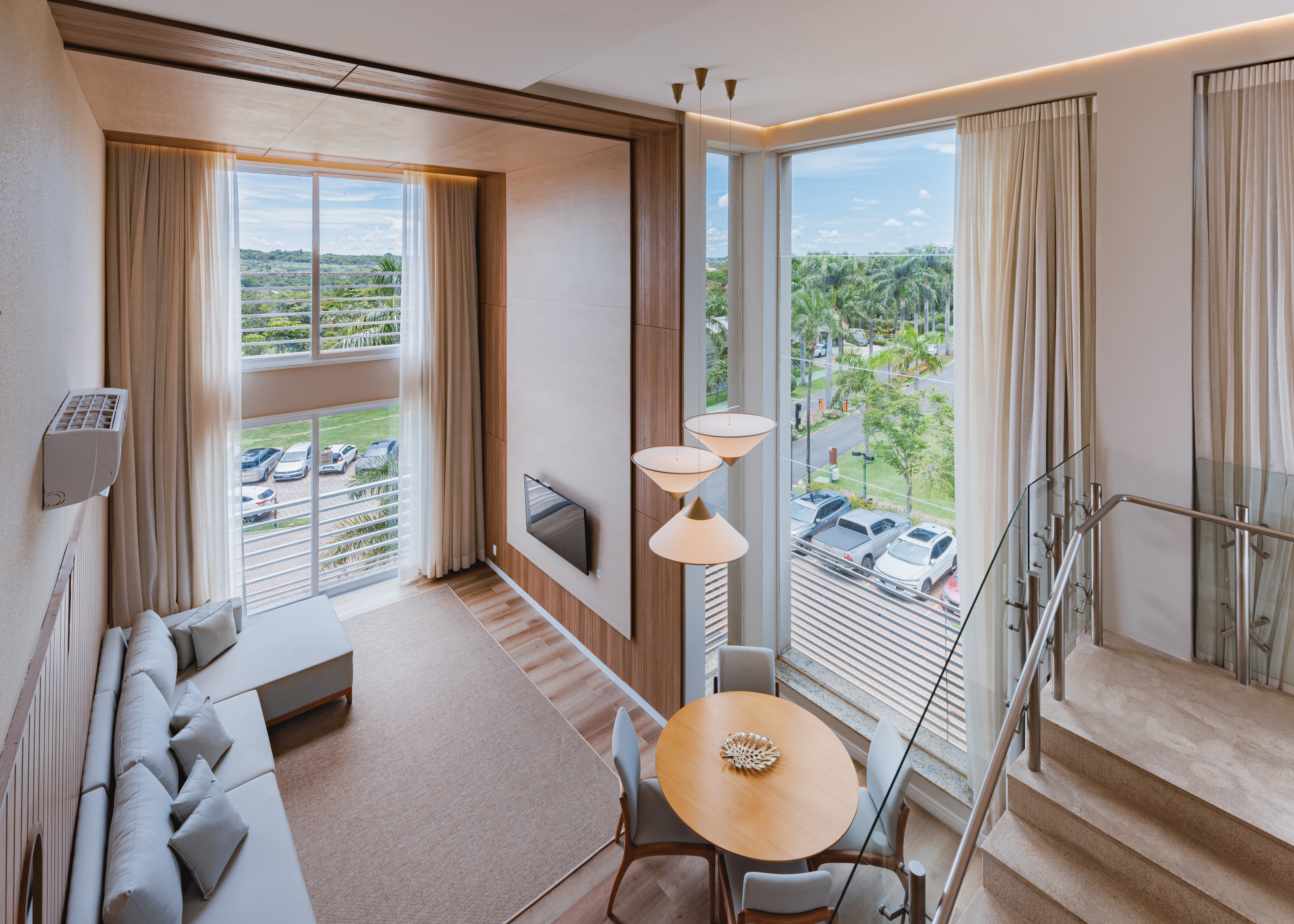 Sala de estar e jantar em um apartamento duplex, com sof&aacute; cinza em L, tapete bege, TV na parede de madeira, mesa de jantar redonda com quatro cadeiras e lumin&aacute;rias pendentes. Grandes janelas com cortinas claras revelam um estacionamento e uma rua arborizada com palmeiras. Uma escada com corrim&atilde;o de vidro e metal aparece &agrave; direita