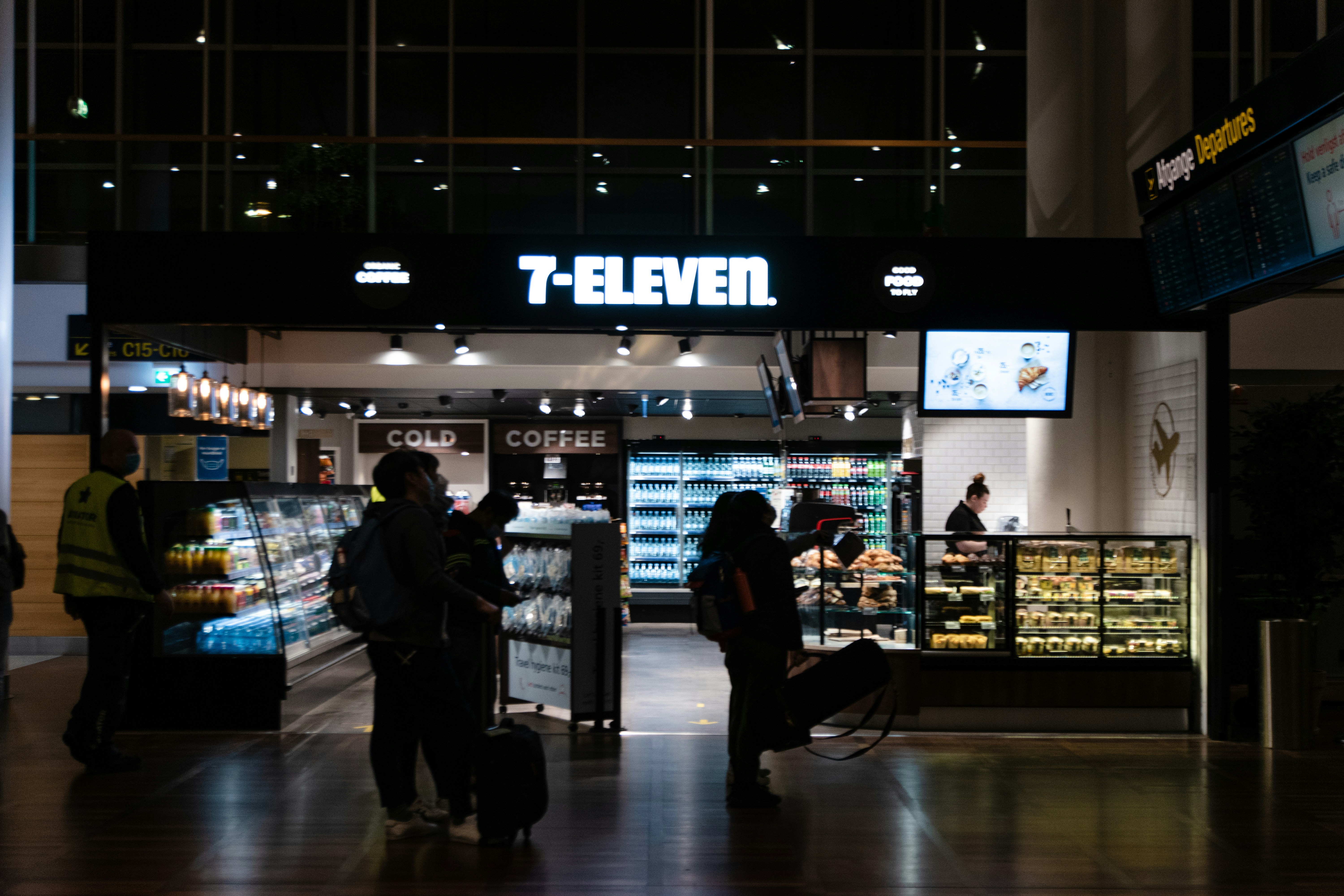 Pessoas em silhueta transitam por um aeroporto escuro, com destaque para uma loja 7-Eleven iluminada ao fundo. Um segurança de colete amarelo e máscara azul caminha à esquerda. À direita, passageiros com malas observam produtos em prateleiras e balcões refrigerados, enquanto uma funcionária atende no caixa. Um letreiro Ankomme Departures aparece no canto superior direito