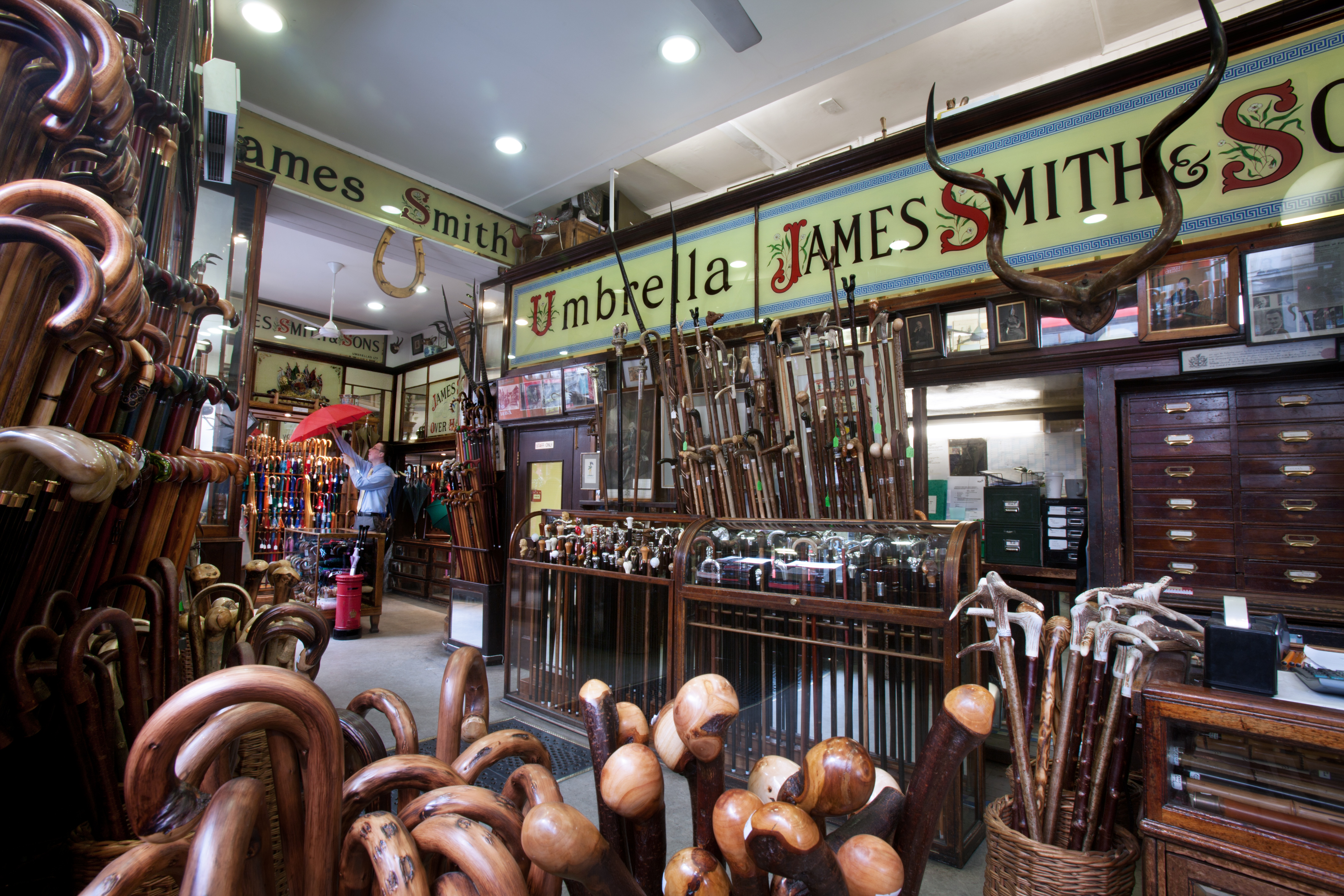 Interior de uma loja antiga e tradicional, James Smith & Sons, especializada em bengalas e guarda-chuvas. Um homem de camisa azul segura um guarda-chuva vermelho aberto, cercado por centenas de bengalas de madeira com diferentes cabos e designs, expostas em cestos e estantes. O ambiente tem iluminação quente, móveis de madeira escura e letreiros vintage, com um par de chifres de touro decorando a parede acima da entrada