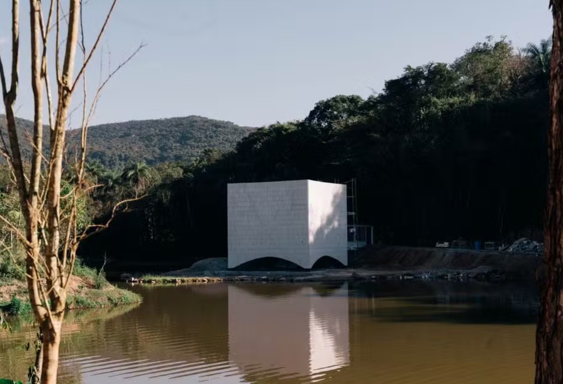 Edifício branco em formato de cubo com base arqueada, refletido em um lago de água escura. Ao fundo, uma floresta densa e montanhas sob céu claro. Um tronco de árvore sem folhas aparece à esquerda, e outro à direita.