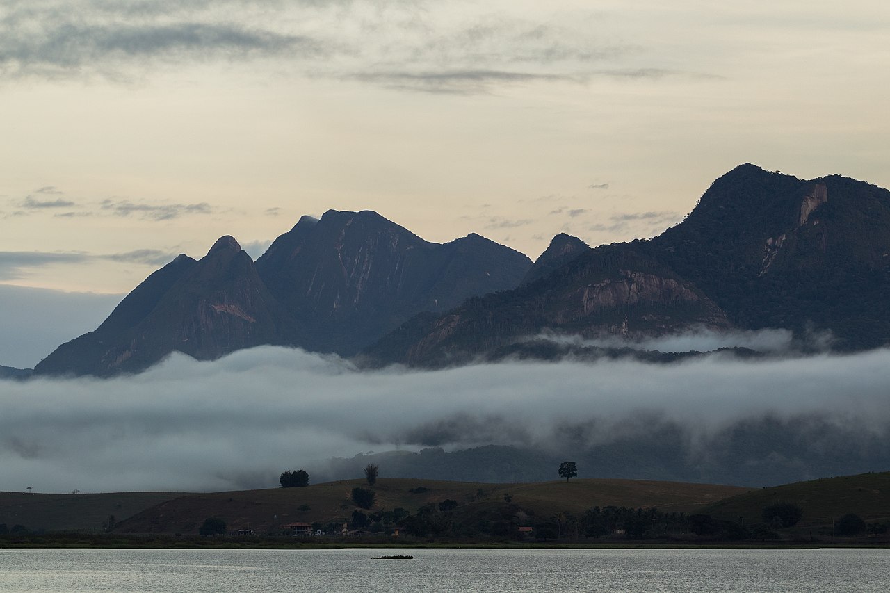Montanhas escuras com picos pontiagudos e vegetação densa, envoltas por uma camada de névoa branca que cobre a base. Abaixo, um lago calmo reflete o céu nublado, com algumas árvores e casas distantes na margem. O céu está cinzento, com nuvens esparsas, sugerindo o amanhecer ou entardecer
