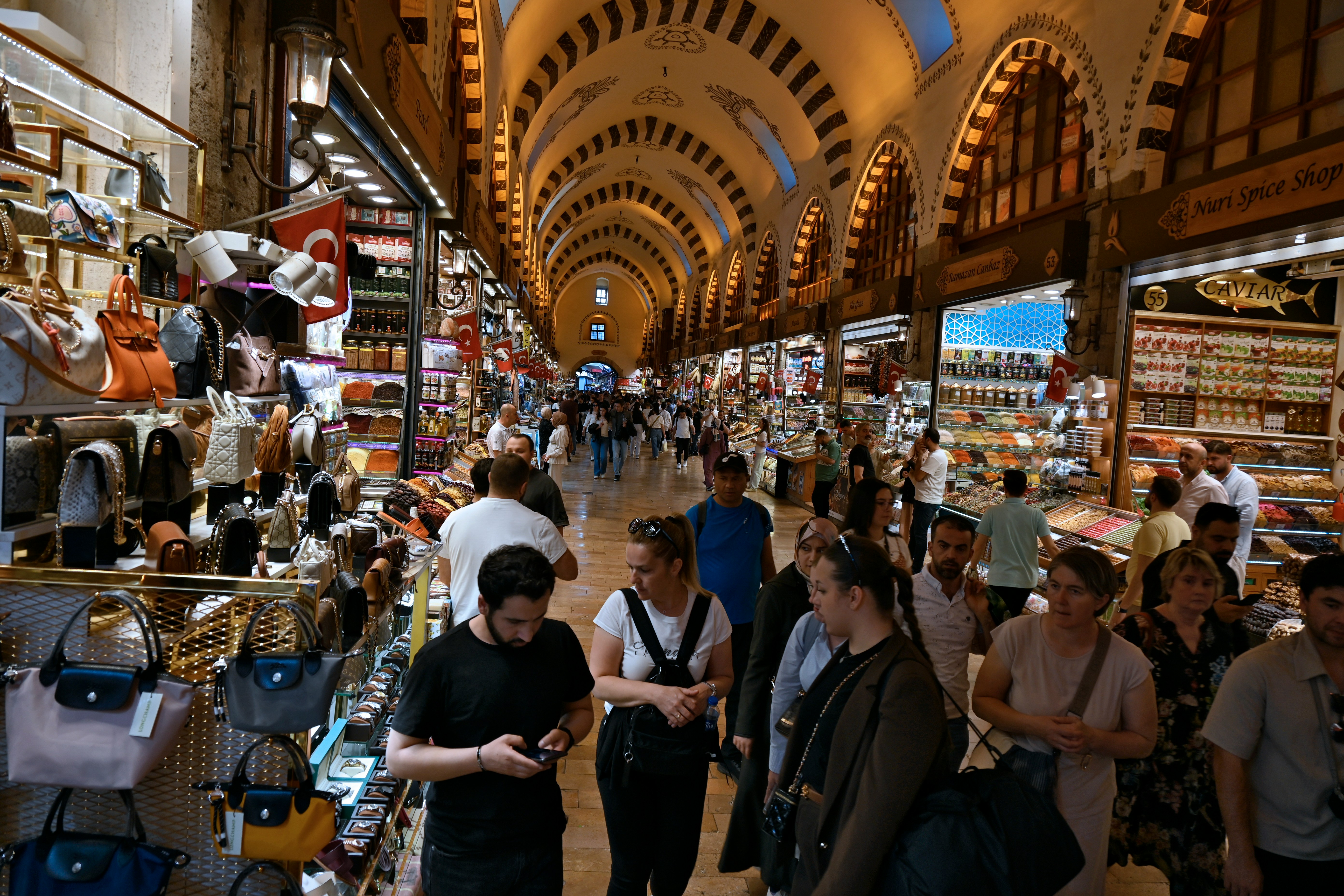 Pessoas caminham por um mercado coberto com teto abobadado e arcos decorados, ladeado por lojas de bolsas, especiarias e doces. Um homem de camiseta preta olha o celular em primeiro plano, enquanto outros clientes e vendedores interagem no ambiente movimentado