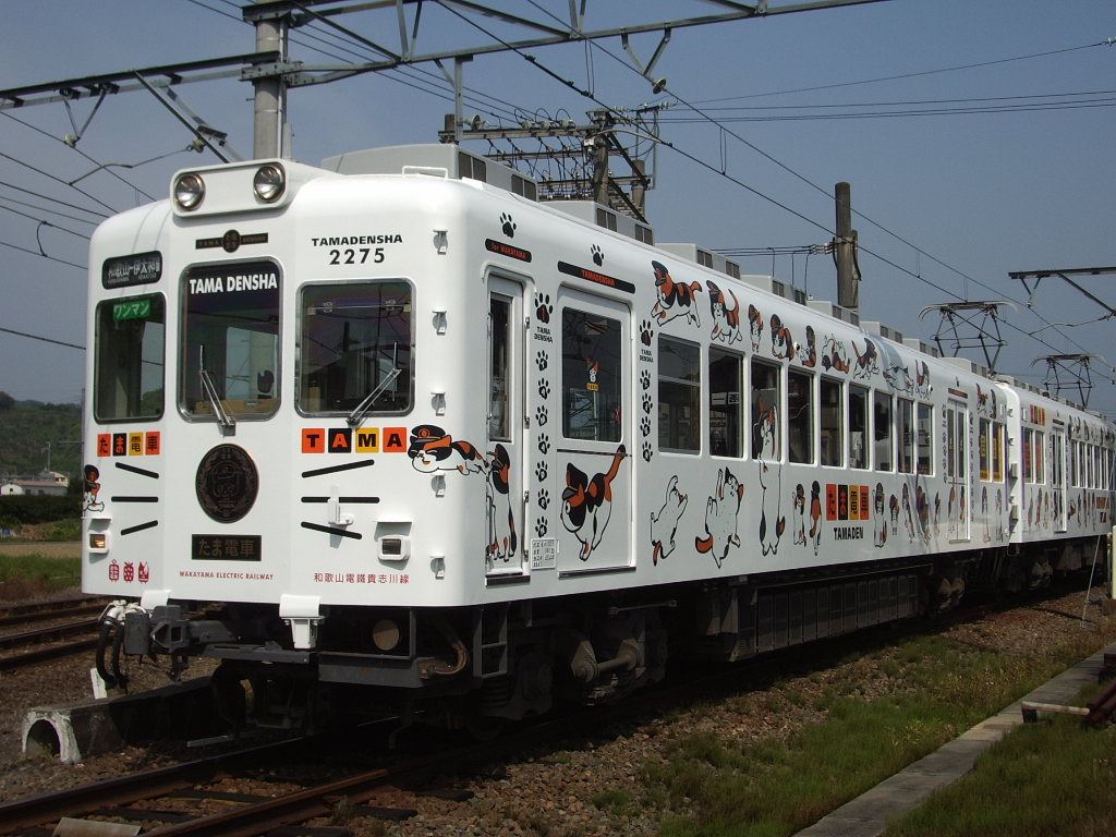 Trem branco com desenhos de gatos e patas pretas e laranjas, parado em trilhos de trem com grama ao lado, sob céu azul claro. Na frente, lê-se TAMA DENSHA 2275 e TAMA em letras laranjas, com um gato de chapéu de maquinista. O trem tem várias janelas e está conectado a fios elétricos aéreos