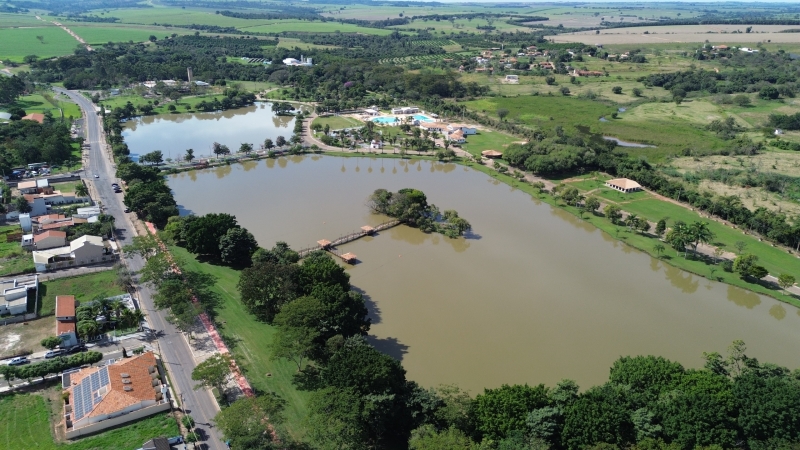 Vista aérea de uma cidade com um grande lago de água barrenta no centro, margeado por uma estrada, casas e árvores. No lago, uma pequena ilha com vegetação e uma passarela de madeira conectando-a à margem. Ao fundo, campos verdes e mais vegetação. À direita do lago, um complexo de lazer com piscinas azuis e edifícios brancos. À esquerda, casas com telhados de telha e painéis solares. O céu está claro com poucas nuvens