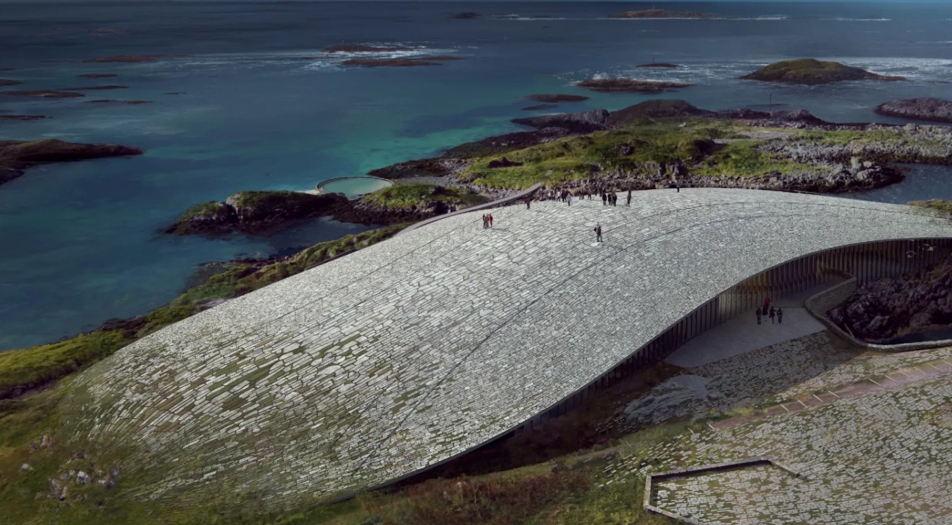 Vista aérea de um edifício moderno com telhado curvo de pedra, integrando-se à paisagem costeira rochosa. Pessoas caminham sobre o telhado e na entrada, com o mar azul-turquesa e ilhas ao fundo