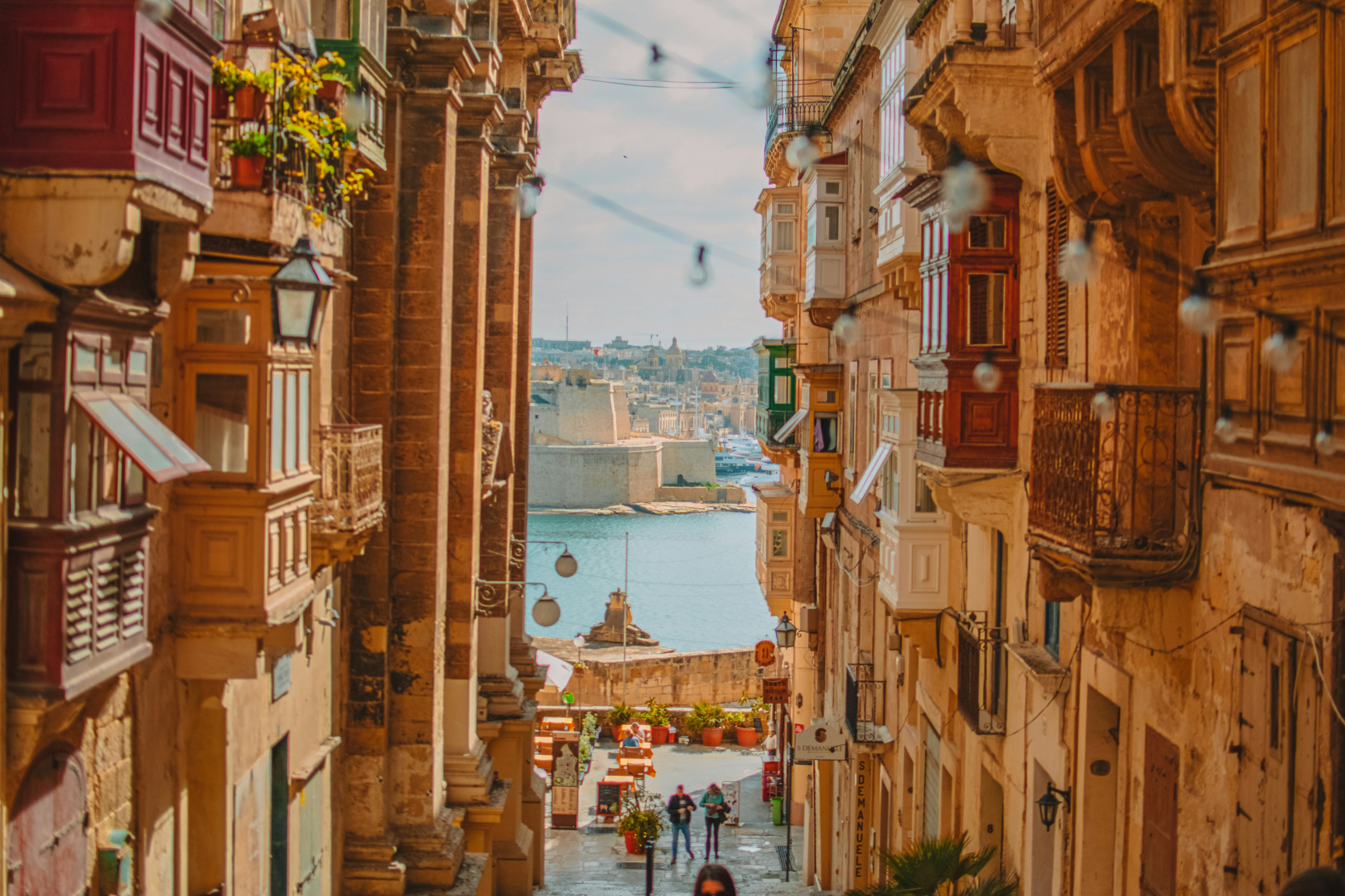 Rua estreita com edifícios antigos de pedra em tons terrosos, varandas coloridas e sacadas de ferro. Ao fundo, o mar azul com barcos e uma fortificação histórica sob céu claro. Pessoas caminham pela rua, e mesas de café estão visíveis.