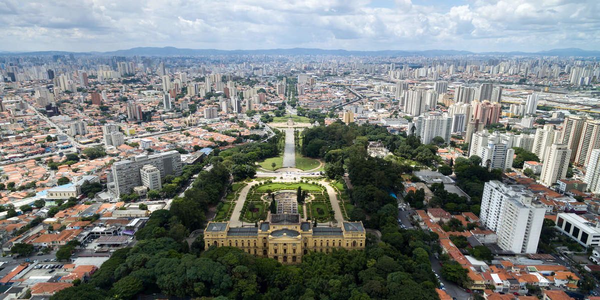 Vista a&eacute;rea do Museu do Ipiranga em S&atilde;o Paulo, um edif&iacute;cio amarelo neocl&aacute;ssico cercado por jardins verdes e uma longa avenida com espelho d&aacute;gua, em contraste com a vasta paisagem urbana de pr&eacute;dios altos e casas com telhados alaranjados sob um c&eacute;u nublado