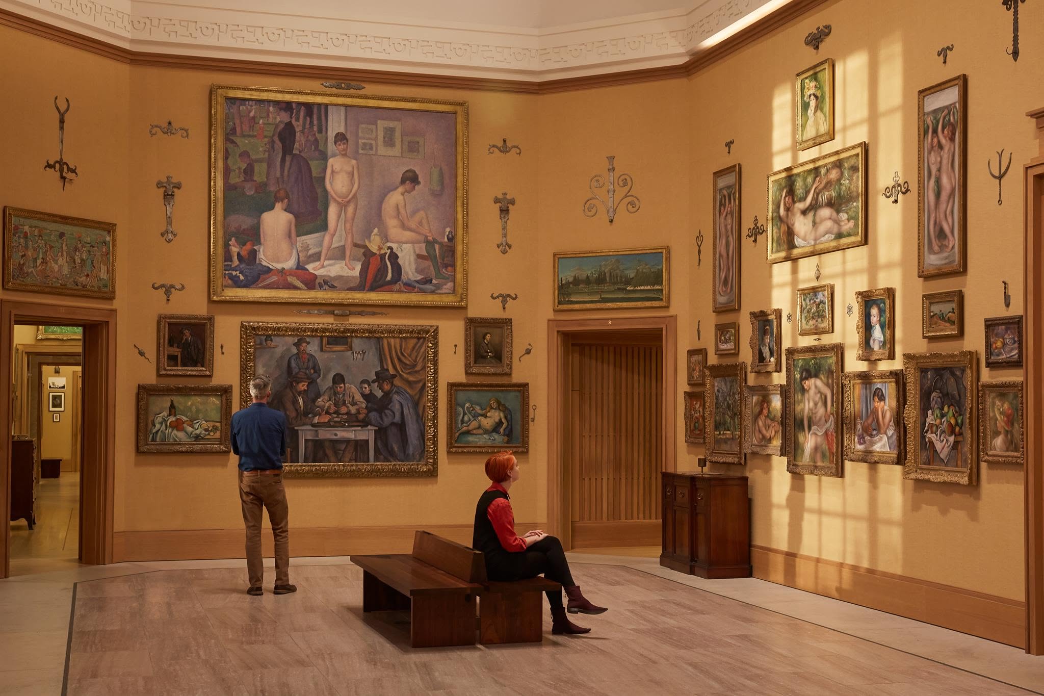 Visitantes em uma galeria de arte com paredes amarelas repletas de quadros emoldurados, incluindo retratos e cenas com figuras nuas. Um homem de camisa azul e calça marrom observa uma pintura de homens jogando cartas, enquanto uma mulher de cabelo ruivo e blusa vermelha senta em um banco de madeira, olhando para as obras. A luz do sol entra pela direita, criando sombras nas paredes