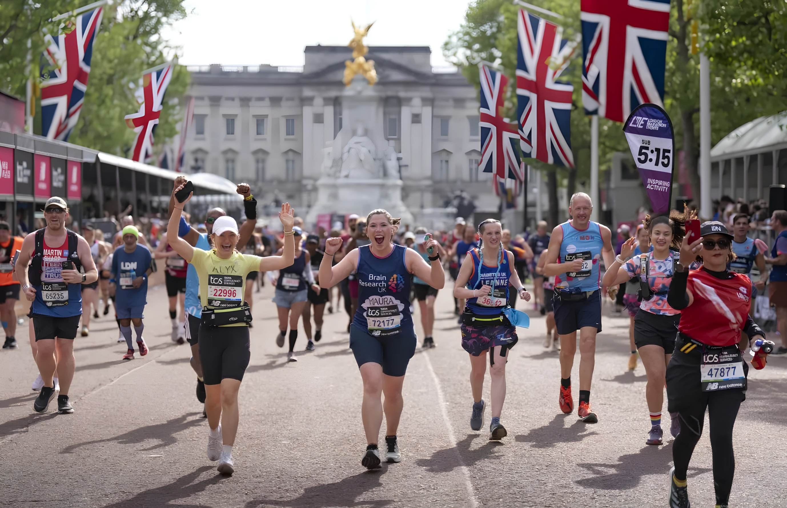 Corredores sorridentes e animados participam de uma maratona em Londres, com o Palácio de Buckingham ao fundo e bandeiras do Reino Unido adornando o percurso
