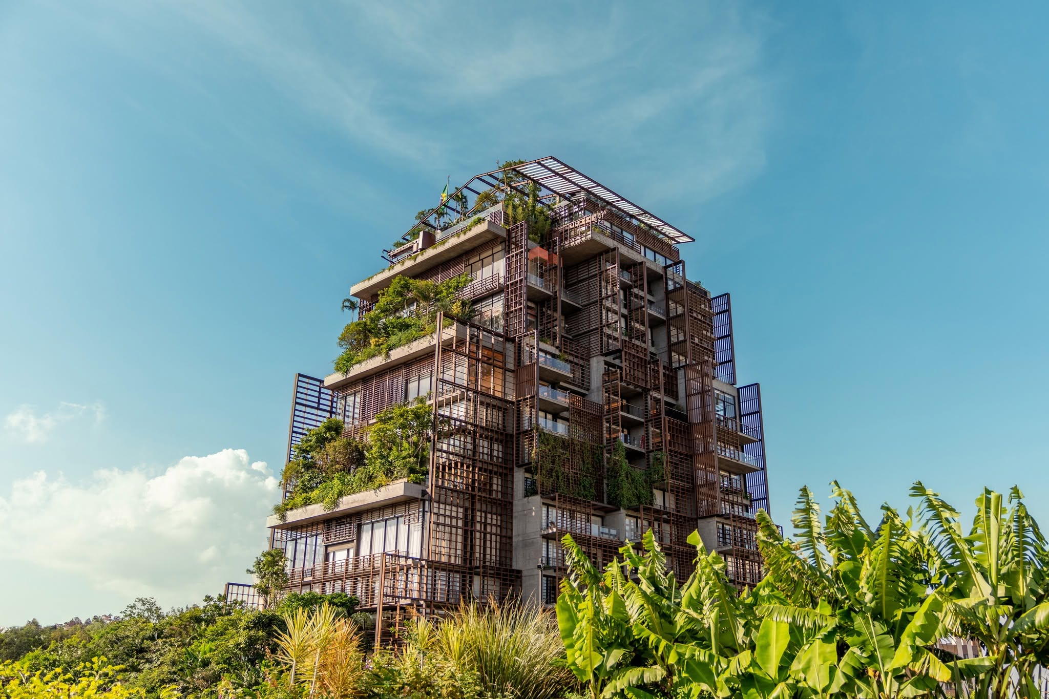 Edifício moderno com varandas e telhado verde, coberto por vegetação exuberante, em um dia ensolarado com céu azul. Plantas tropicais em primeiro plano.