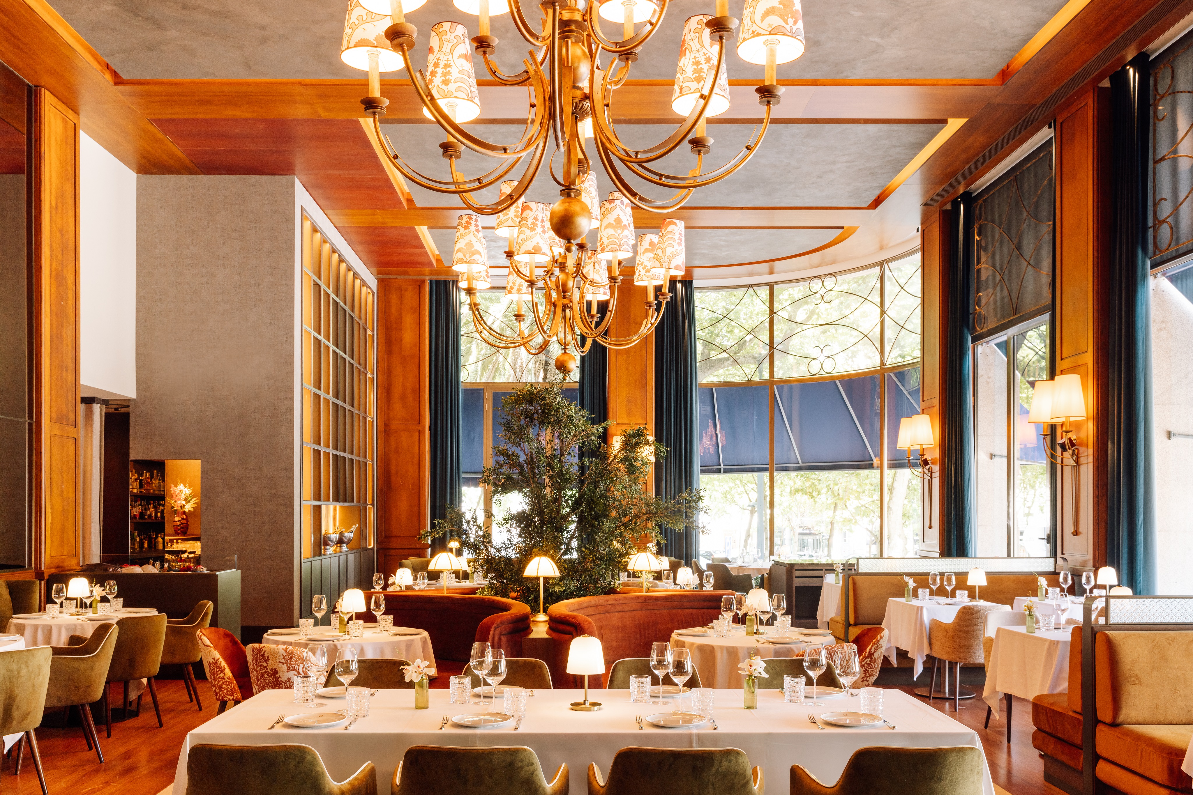 Interior de um restaurante elegante com mesas postas, cadeiras e sofás estofados em tons terrosos. Um grande lustre dourado com cúpulas estampadas ilumina o ambiente, que possui paredes de madeira e janelas amplas com cortinas azuis e detalhes em ferro forjado. Plantas verdes decoram o centro do salão, criando uma atmosfera sofisticada e acolhedora