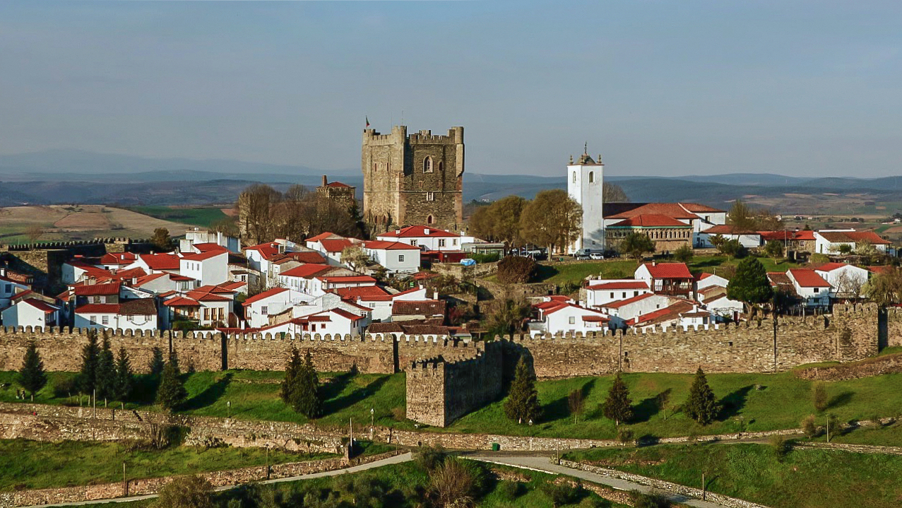 Vista aérea de uma vila histórica murada, com casas brancas de telhados vermelhos. Ao fundo, um castelo medieval de pedra e uma igreja branca com torre sineira. A muralha de pedra envolve a vila, e o entorno é de colinas verdes e campos. O céu é azul claro, indicando um dia ensolarado