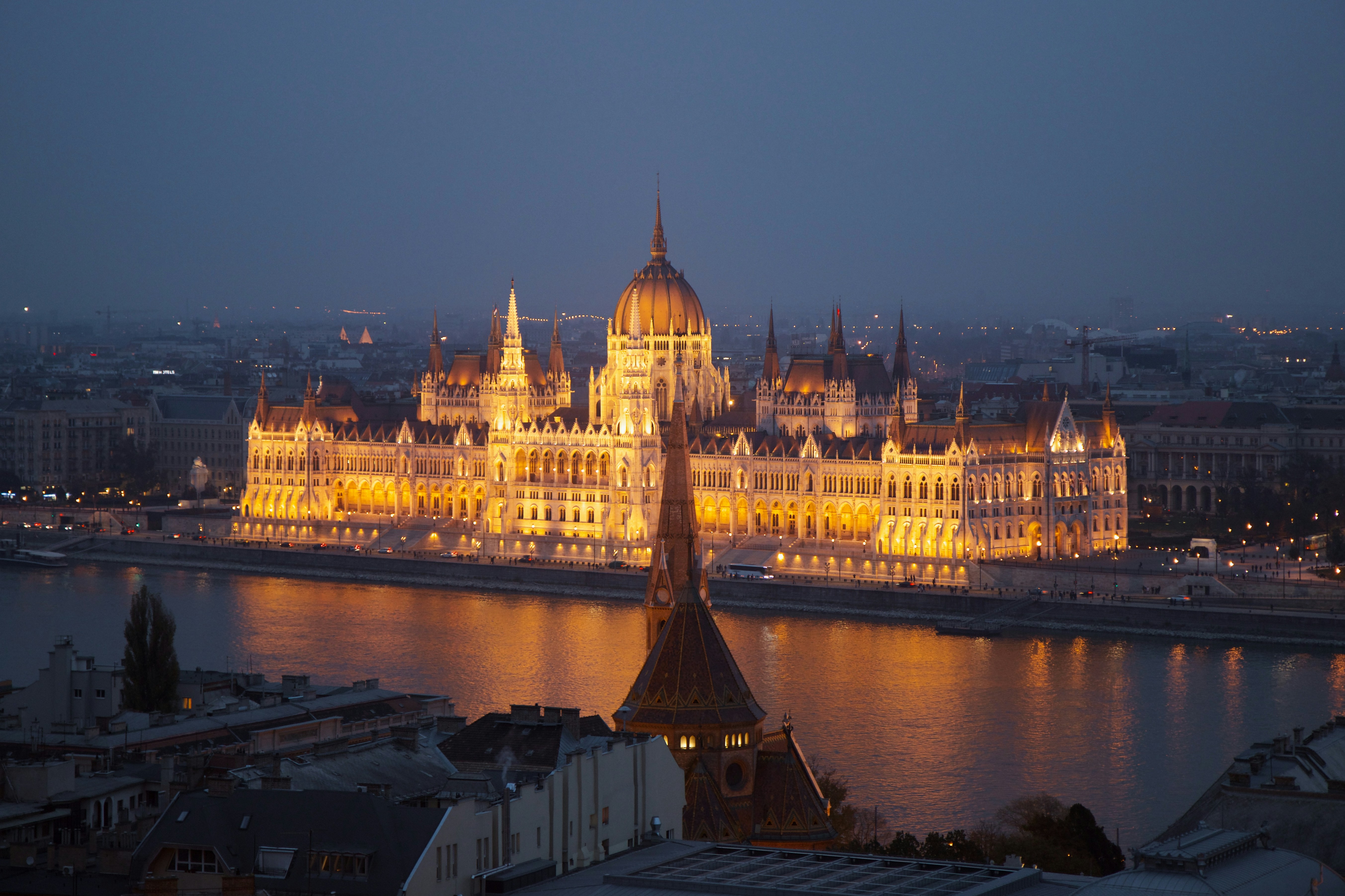O Parlamento Húngaro iluminado em amarelo-ouro à noite, com sua cúpula central e torres pontiagudas, refletindo na água escura do rio Danúbio. Prédios escuros e uma torre pontiaguda em primeiro plano, com a cidade ao fundo sob um céu azul-escuro.