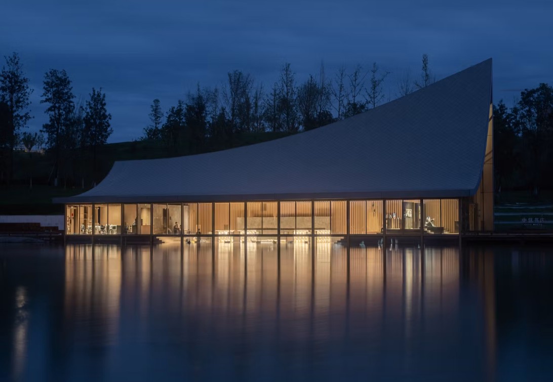 Edifício moderno com telhado curvo e inclinado, janelas de vidro iluminadas, refletindo na água escura de um lago ao anoitecer. Árvores escuras no fundo.