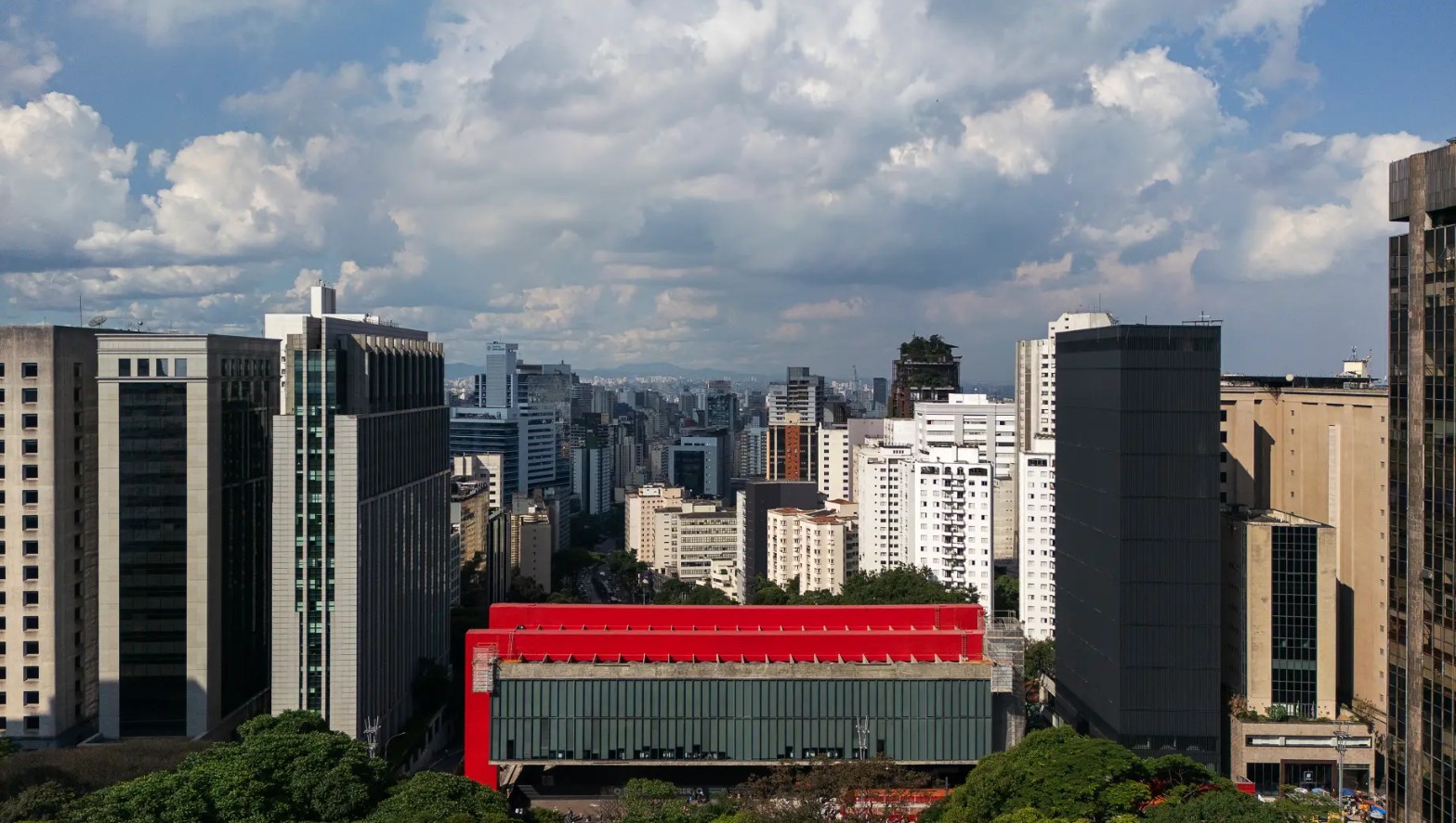 Vista aérea de uma metrópole com edifícios altos e densos sob um céu azul com nuvens brancas. Em primeiro plano, o MASP com sua estrutura vermelha e fachada de vidro verde, cercado por árvores.