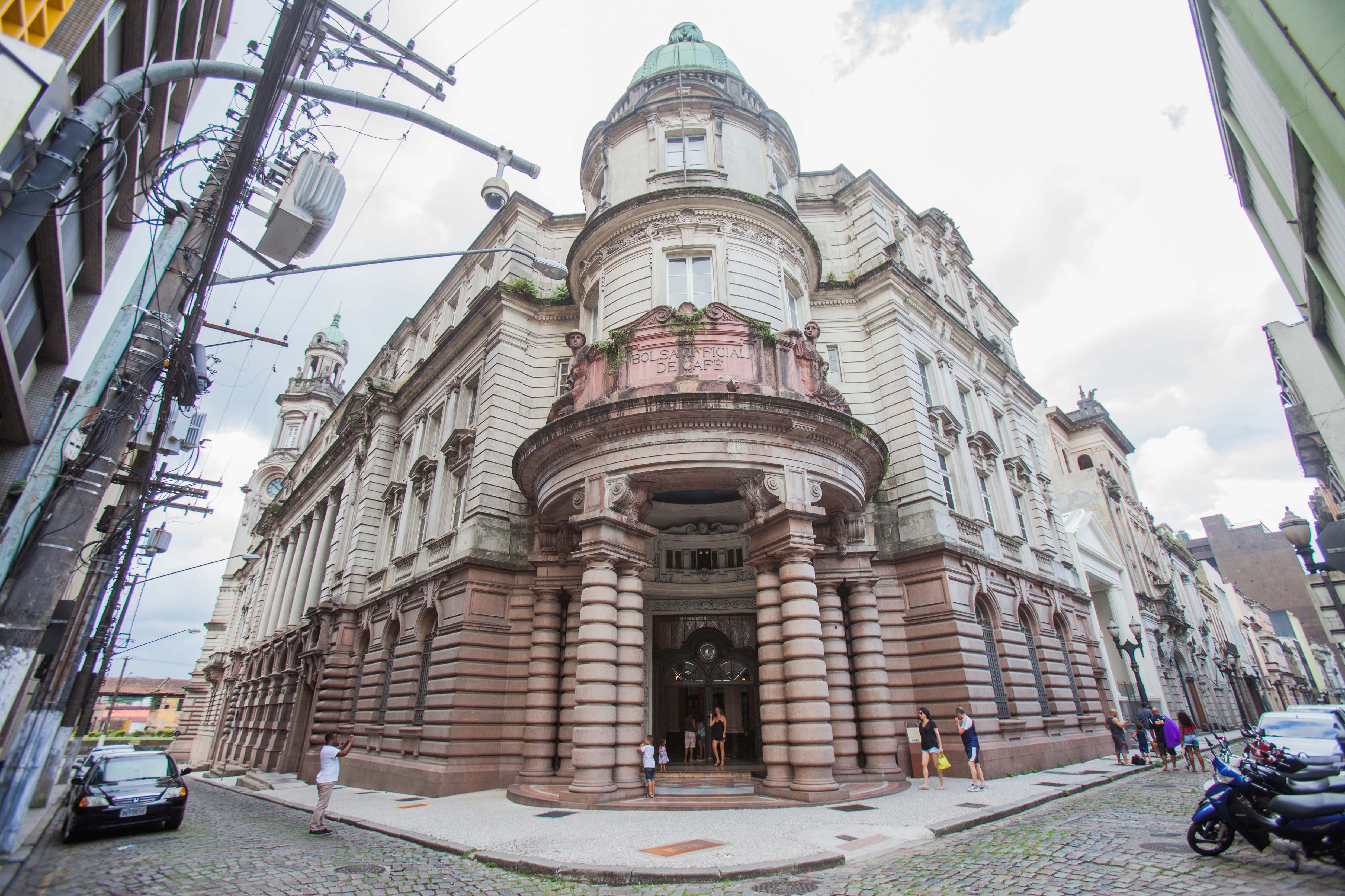 Fachada da Bolsa Oficial de Café em Santos, edifício histórico com cúpula verde, varanda rosa com a inscrição Bolsa Official de Café e colunas robustas. Pessoas caminham na rua de paralelepípedos, com postes e fiação elétrica à esquerda e carros estacionados