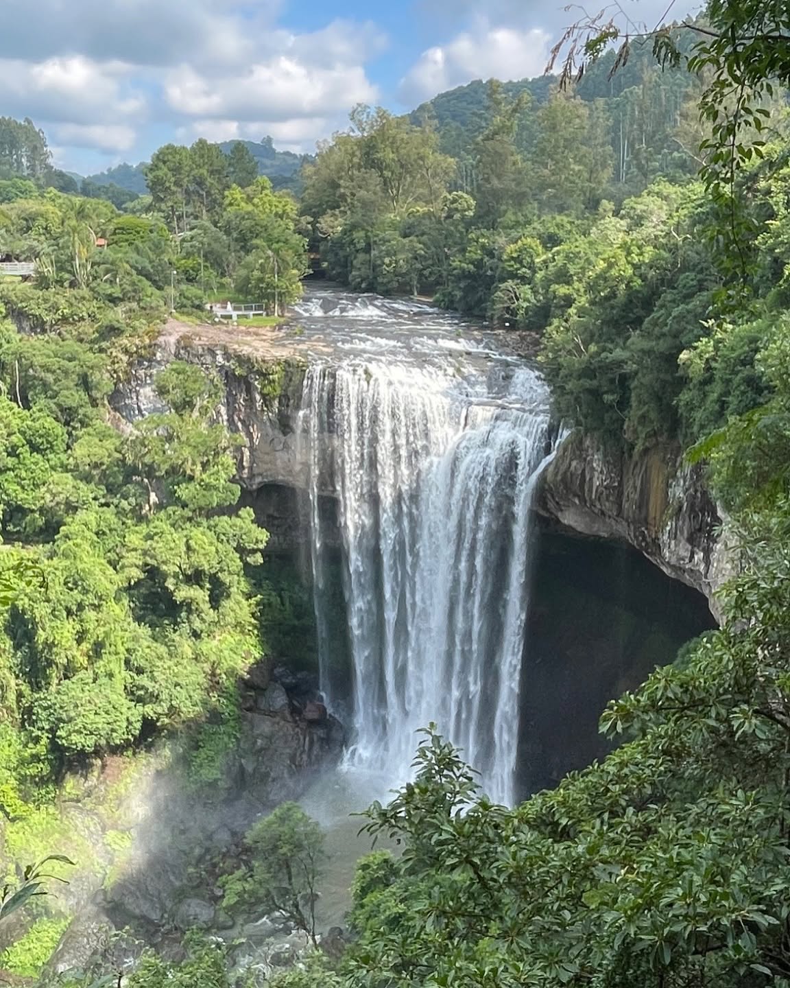 Cachoeira imponente com grande volume de água caindo de um penhasco rochoso, cercada por densa vegetação verde e montanhas ao fundo, sob céu azul com nuvens esparsas
