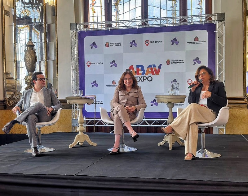 Três pessoas sentadas em cadeiras brancas em um palco escuro, com um painel roxo e branco da ABAV Expo ao fundo. Um homem de óculos e terno cinza está à esquerda, uma mulher de conjunto bege sorri no centro, e outra mulher de blazer preto e calça bege fala ao microfone à direita