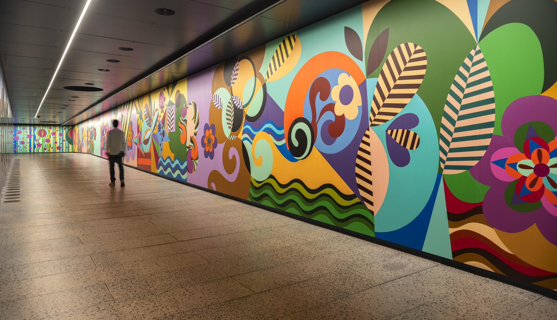 Pessoa de costas caminhando por um corredor iluminado com um mural colorido de formas abstratas e florais na parede direita
